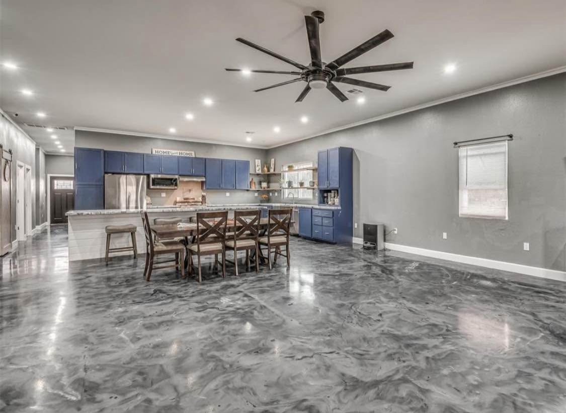 TBC project photo of an open living and dining area with a dining table, blue kitchen cabinetry, recessed lights, and a large ceiling fan.