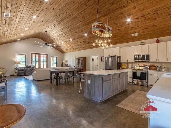 TBC project photo of an open kitchen and living area with white cabinets, a wood island, dining table, polished concrete floor, and wood plank ceiling.