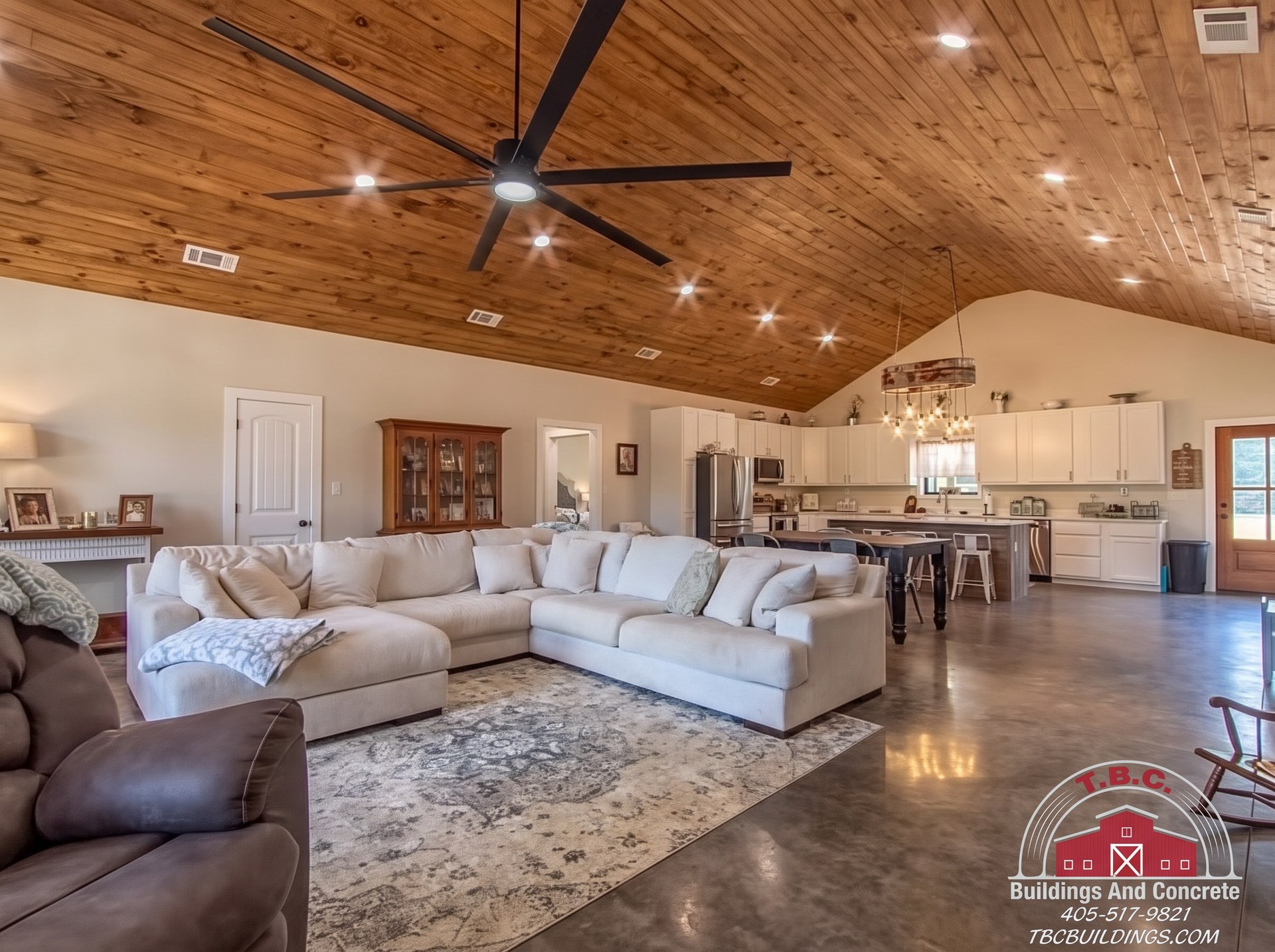 TBC project photo of an open living room and kitchen with sectional seating, white cabinets, polished concrete floor, and wood plank ceiling.