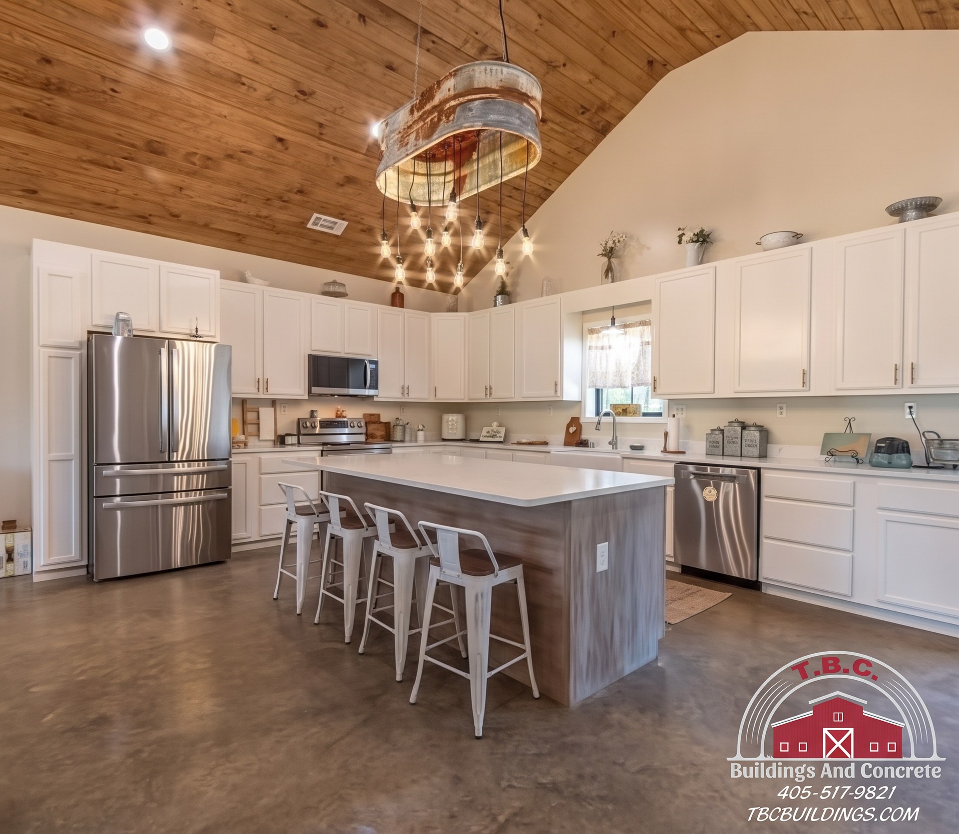 TBC project photo of a kitchen with white cabinets, stainless appliances, a wood island with stools, polished concrete floor, and wood plank ceiling.