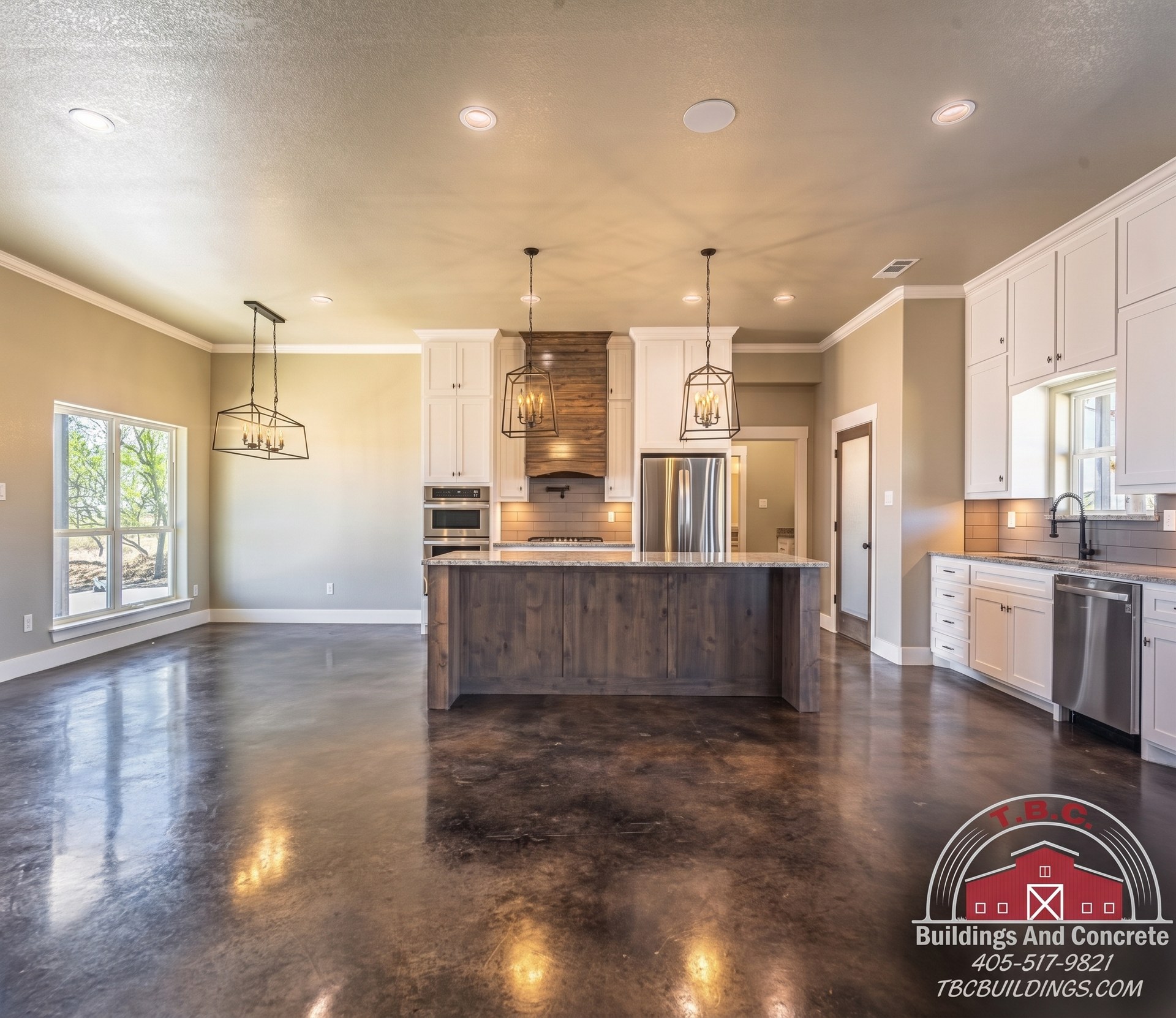 TBC project photo of a kitchen with white cabinets, a dark wood island, pendant lights, stainless appliances, and polished concrete floor.
