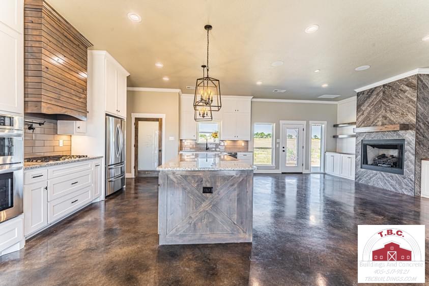 TBC project photo of an open kitchen and living area with white cabinets, a patterned wood island, stone-look fireplace surround, and polished concrete floor.