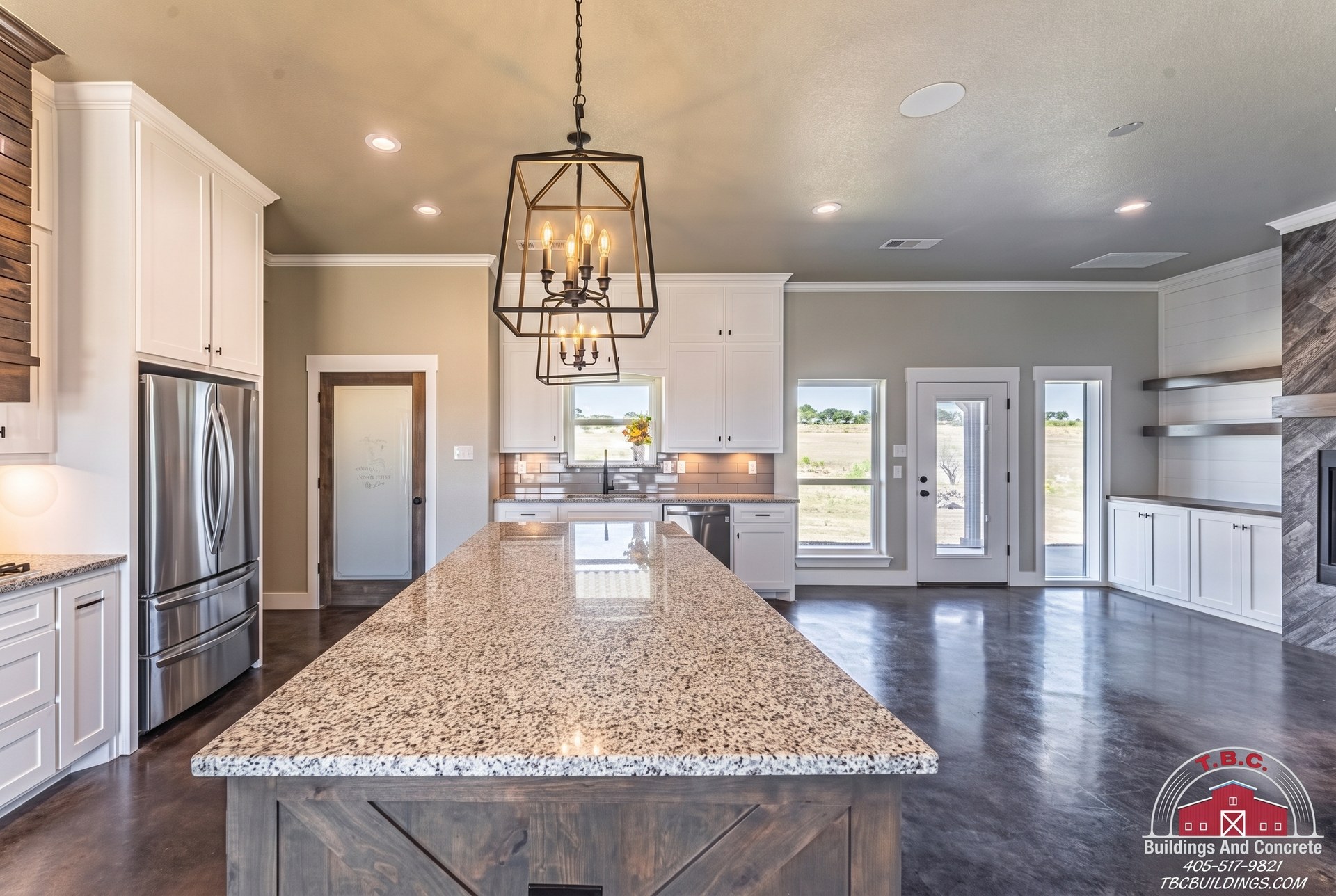 TBC project photo of a kitchen island with a light stone countertop, white cabinets, stainless refrigerator, and adjoining living area.