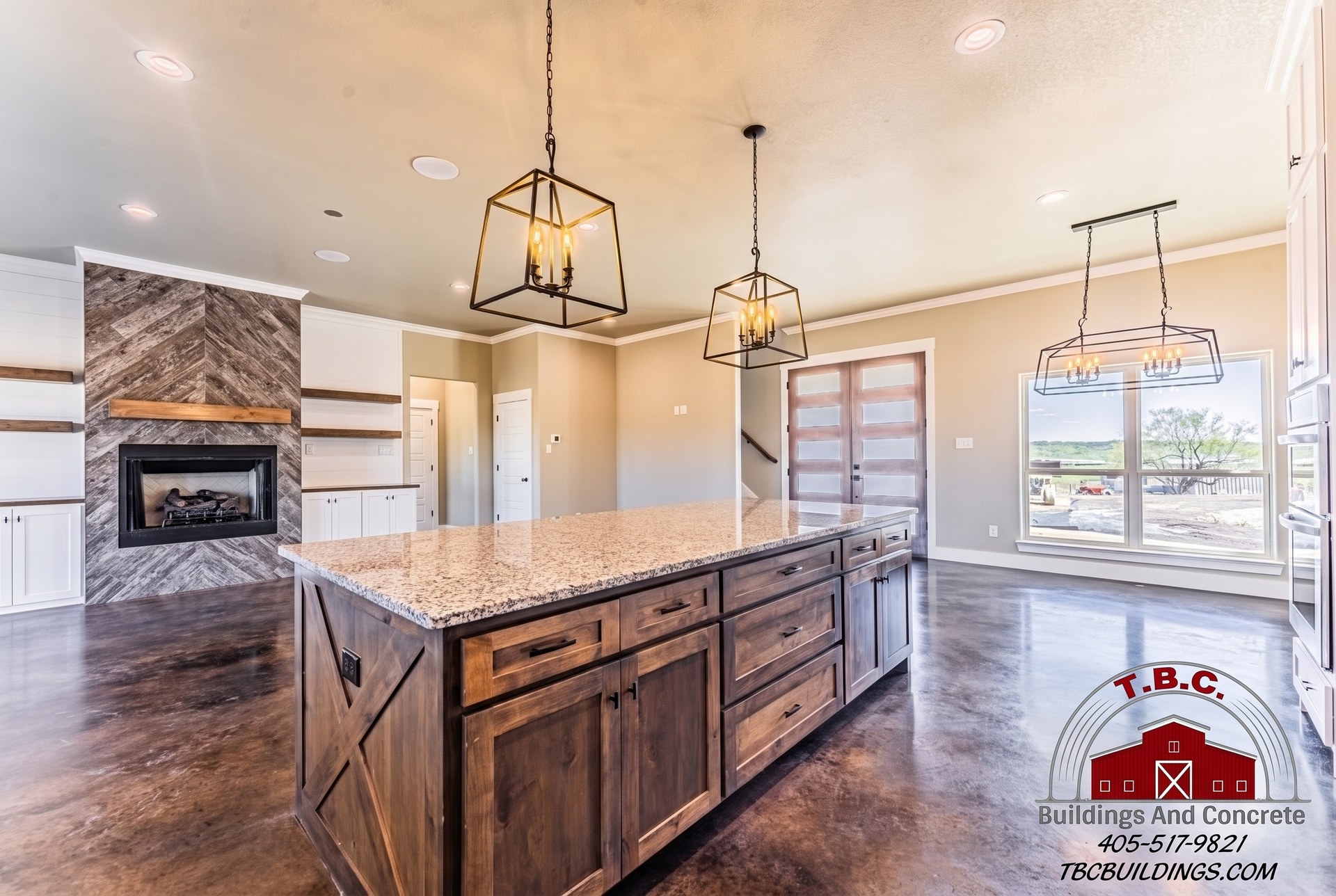 TBC project photo of a wood kitchen island with a light stone countertop, pendant lights, polished concrete floor, and adjoining fireplace wall.
