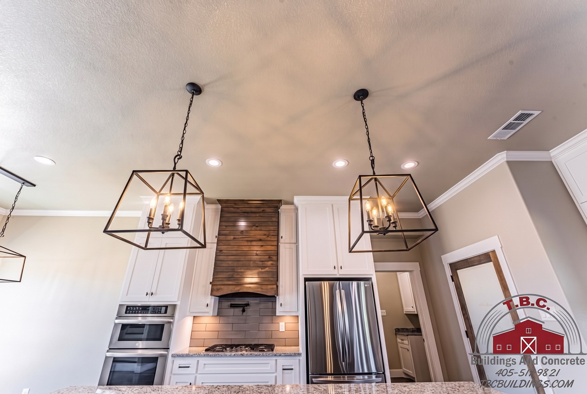 TBC project photo of a finished kitchen with white cabinets, pendant lights, dark island, and adjoining living area.