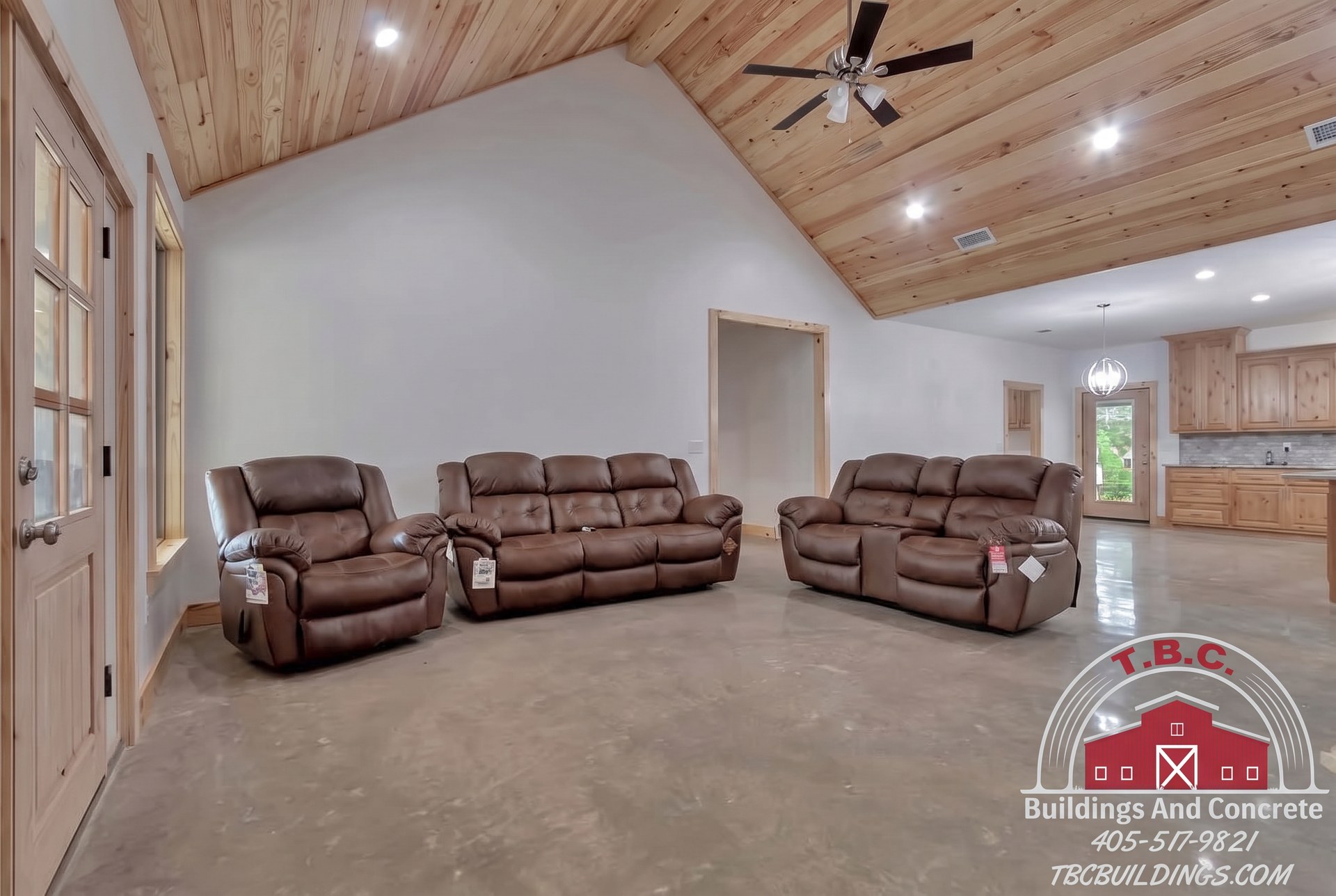TBC project photo of a living area with leather seating, a vaulted wood ceiling, and an adjoining kitchen.