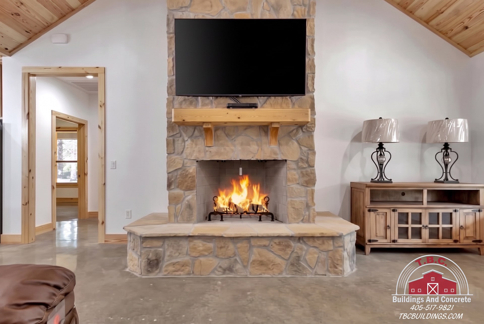 TBC project photo of a stone fireplace with a burning fire, wood mantel, wall-mounted television, and wood-paneled ceiling.