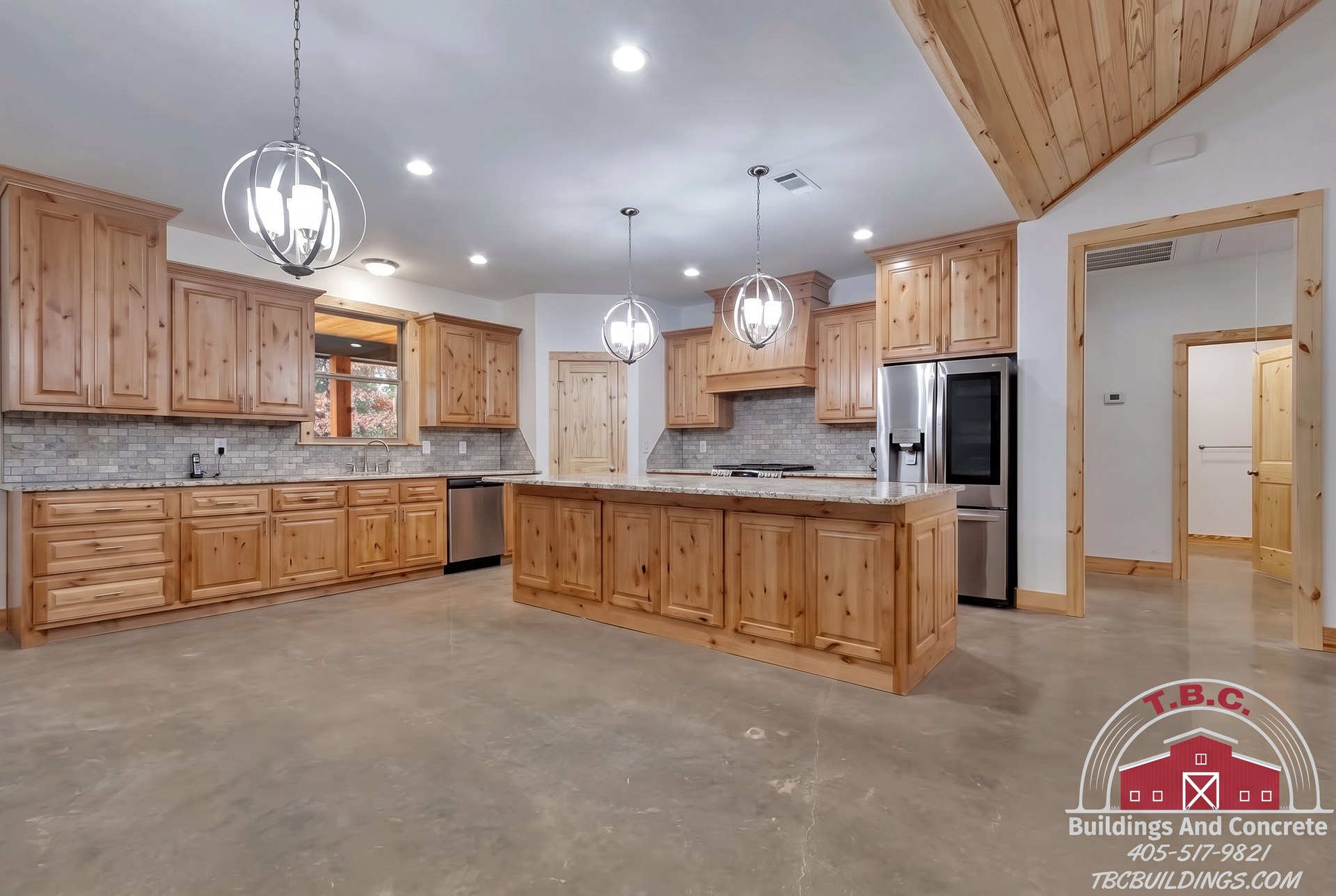 TBC project photo of a kitchen with wood cabinets, a large island, pendant lights, and stainless steel appliances.