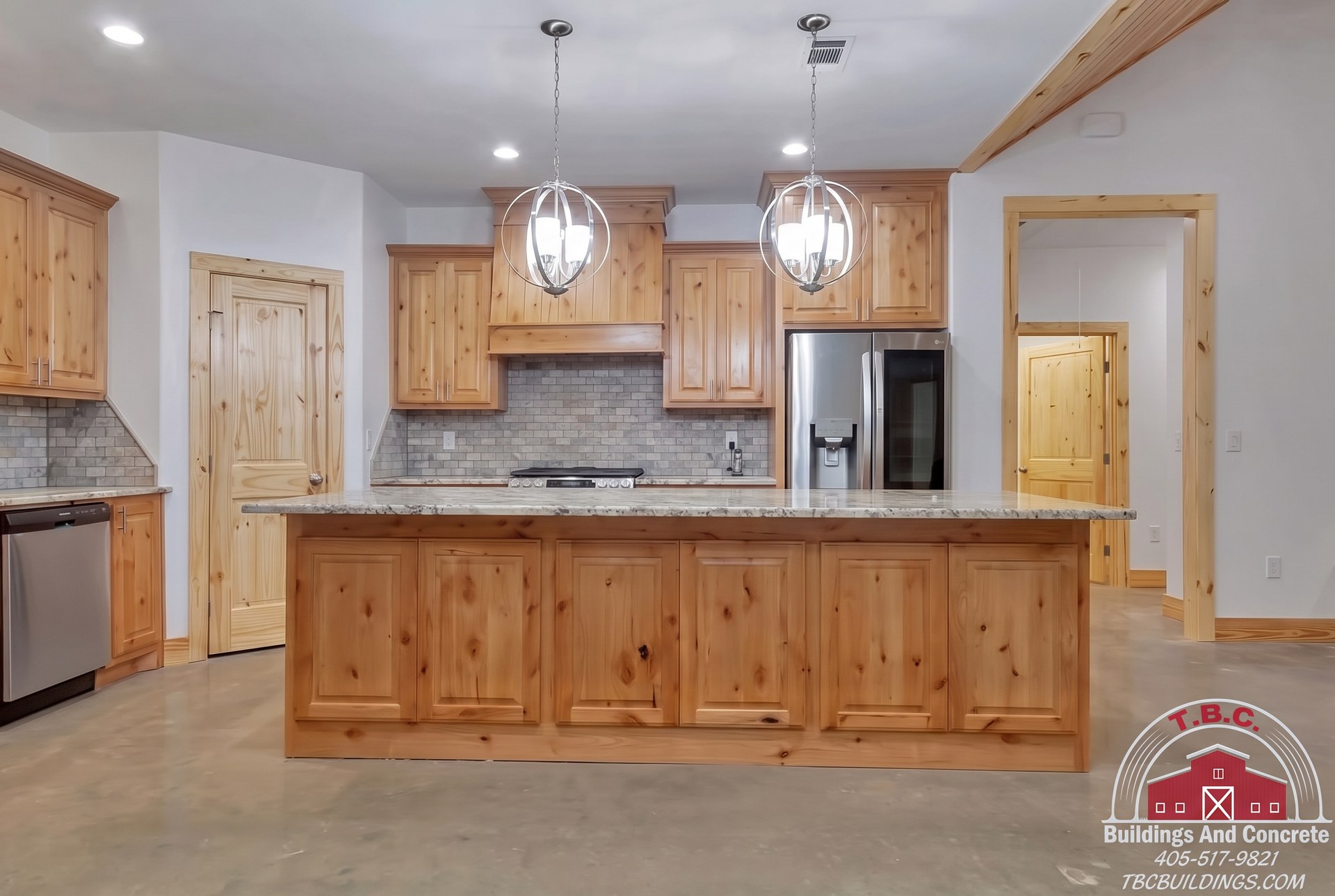 TBC project photo of a kitchen with a wood-front island, pendant lights, wood cabinetry, and polished concrete floor.