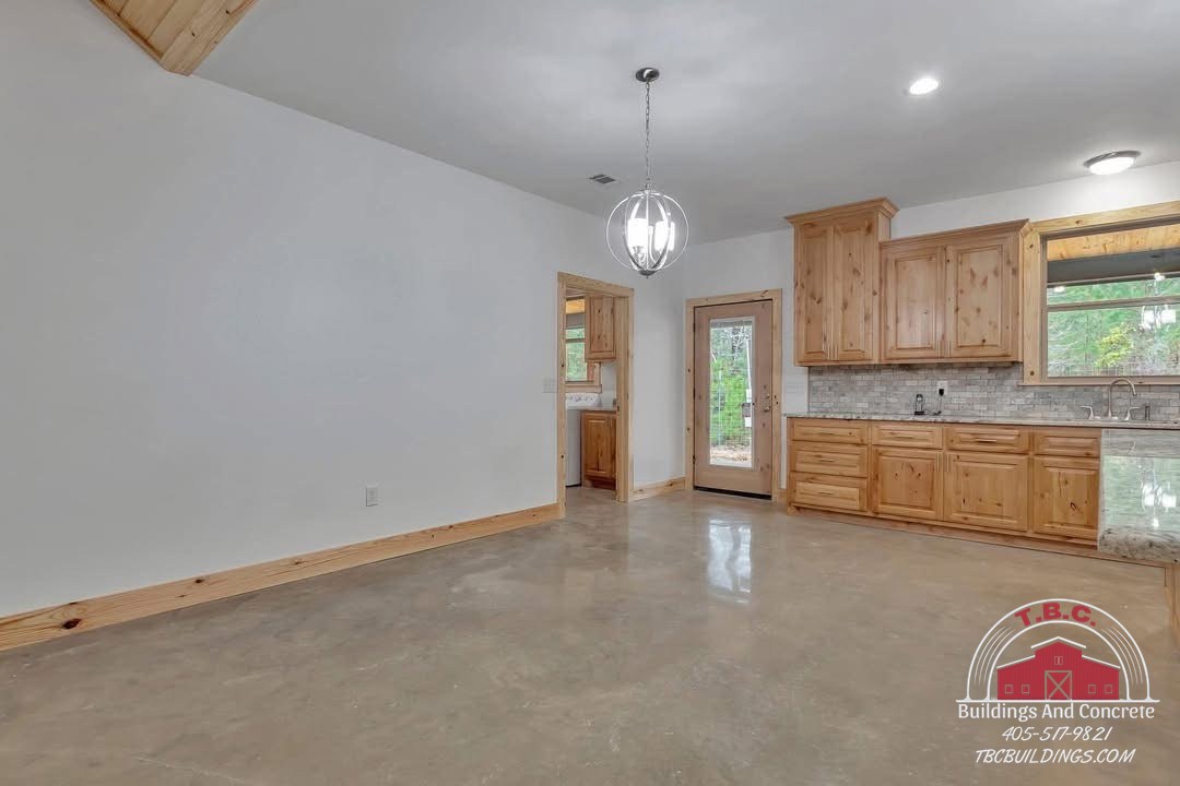 TBC project photo of an open finished room with a wood kitchen wall, pendant light, windows, and polished concrete floor.