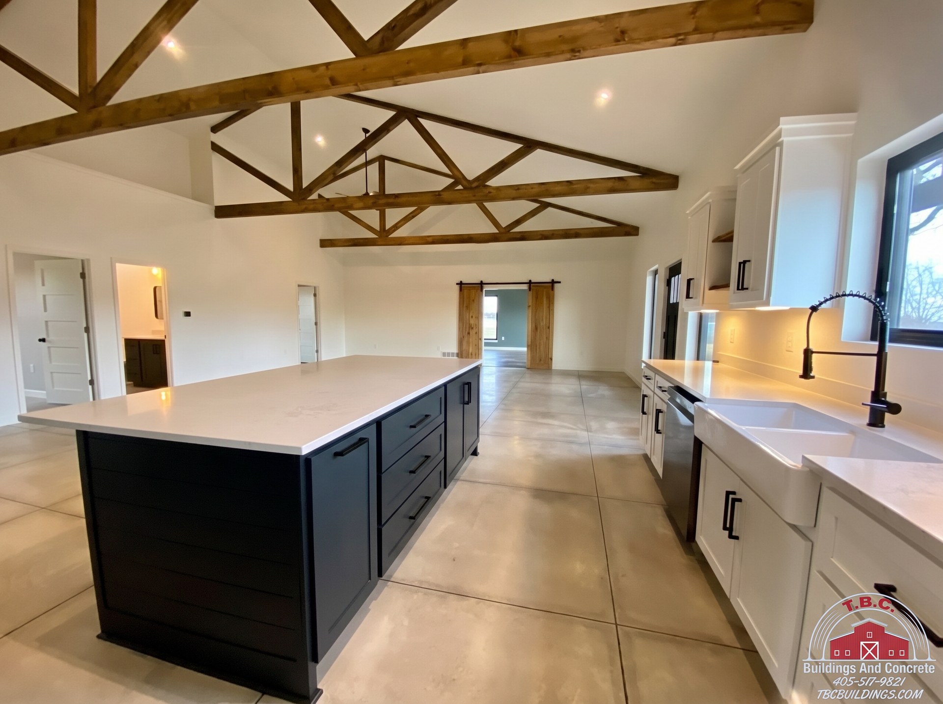 TBC project photo of an open kitchen with a dark island, white cabinetry, exposed wood trusses, and tiled floor.