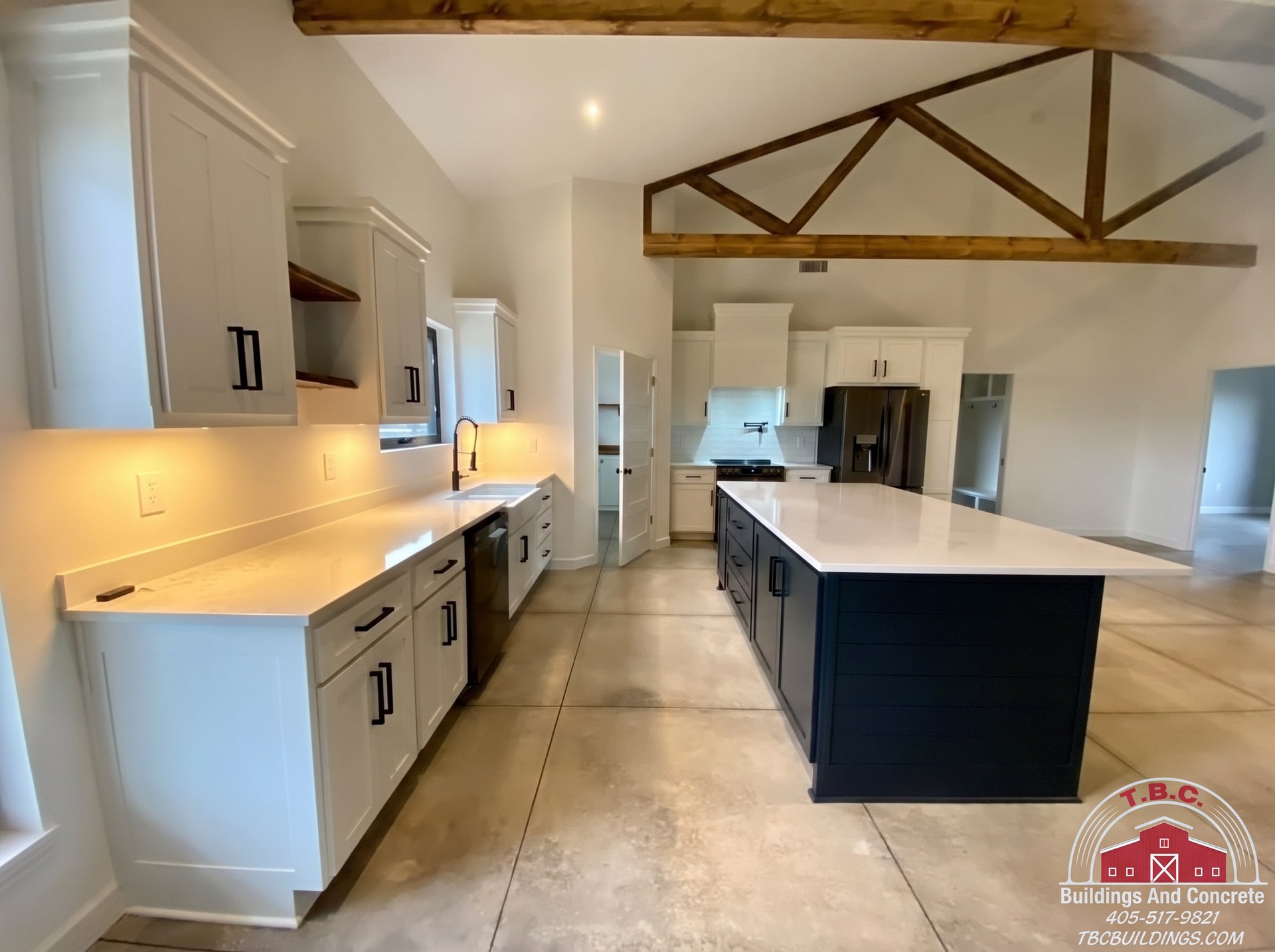 TBC project photo of an open kitchen with a dark island, white cabinets, wood trusses, and tiled floor.