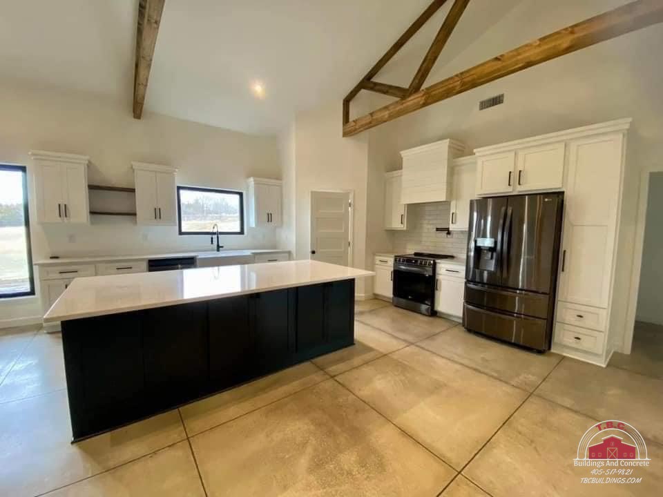 TBC project photo of an open kitchen with white cabinets, a dark island, exposed wood trusses, and tiled floor.