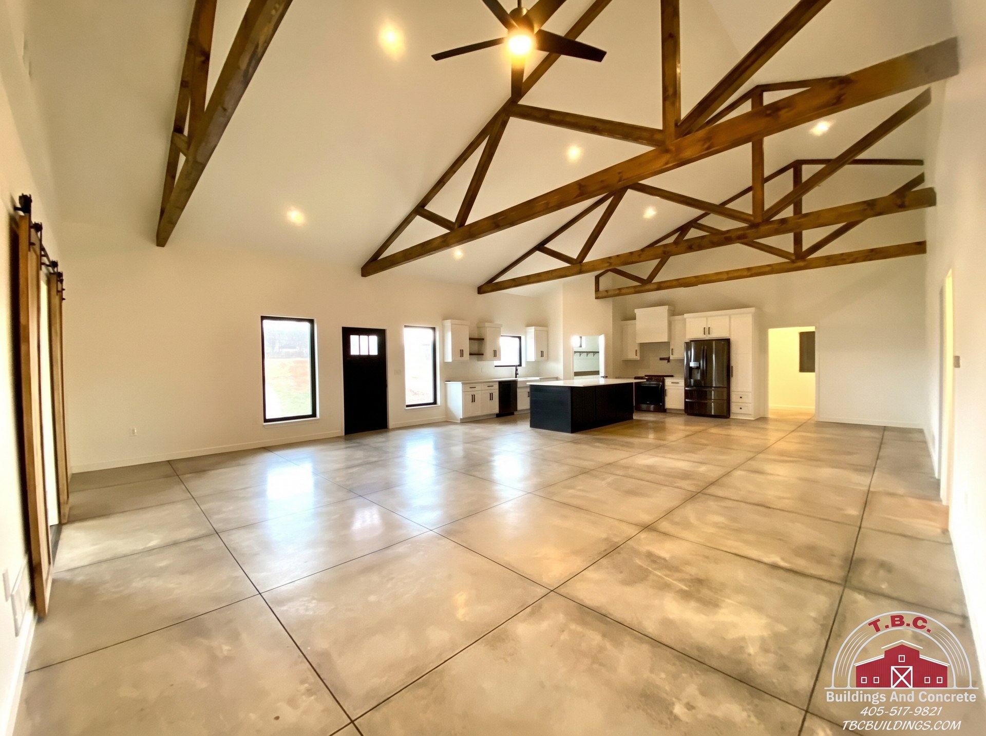 TBC project photo of a large open finished room with exposed wood trusses, tiled floor, ceiling fan, and kitchen in the background.