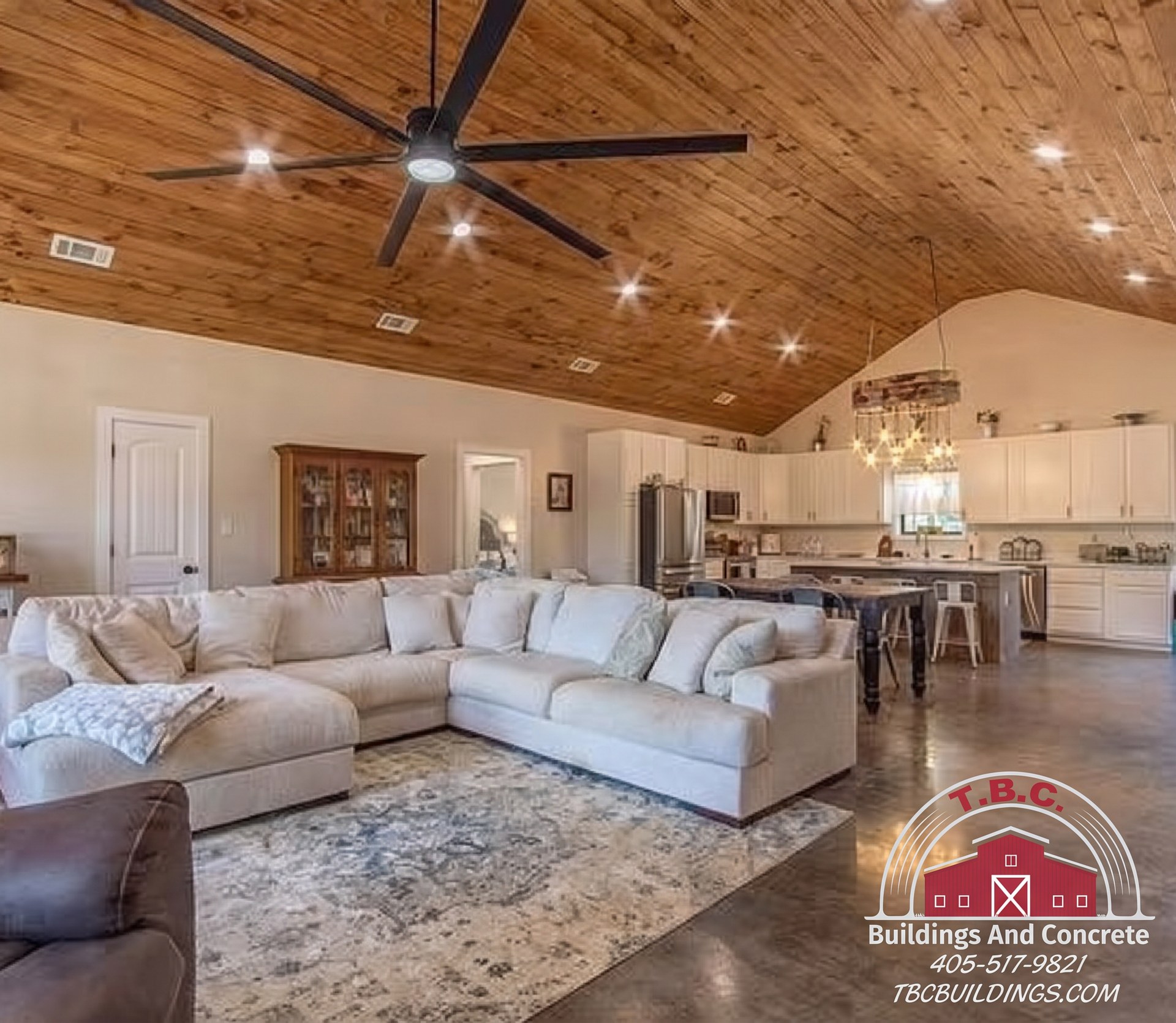 TBC project photo of an open great room with a wood plank ceiling, ceiling fan, sectional seating, and a kitchen in the background.