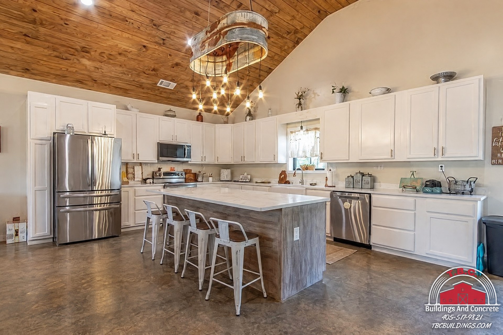 TBC project photo of a white kitchen with a gray island, stainless steel appliances, bar stools, and a wood plank ceiling.