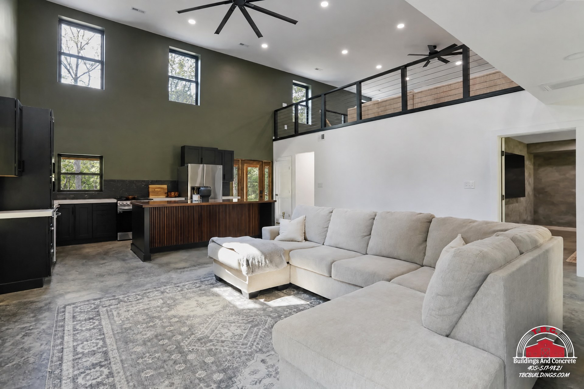 TBC project photo of a two-story great room with a loft railing, sectional seating, a black fireplace wall, and high windows.