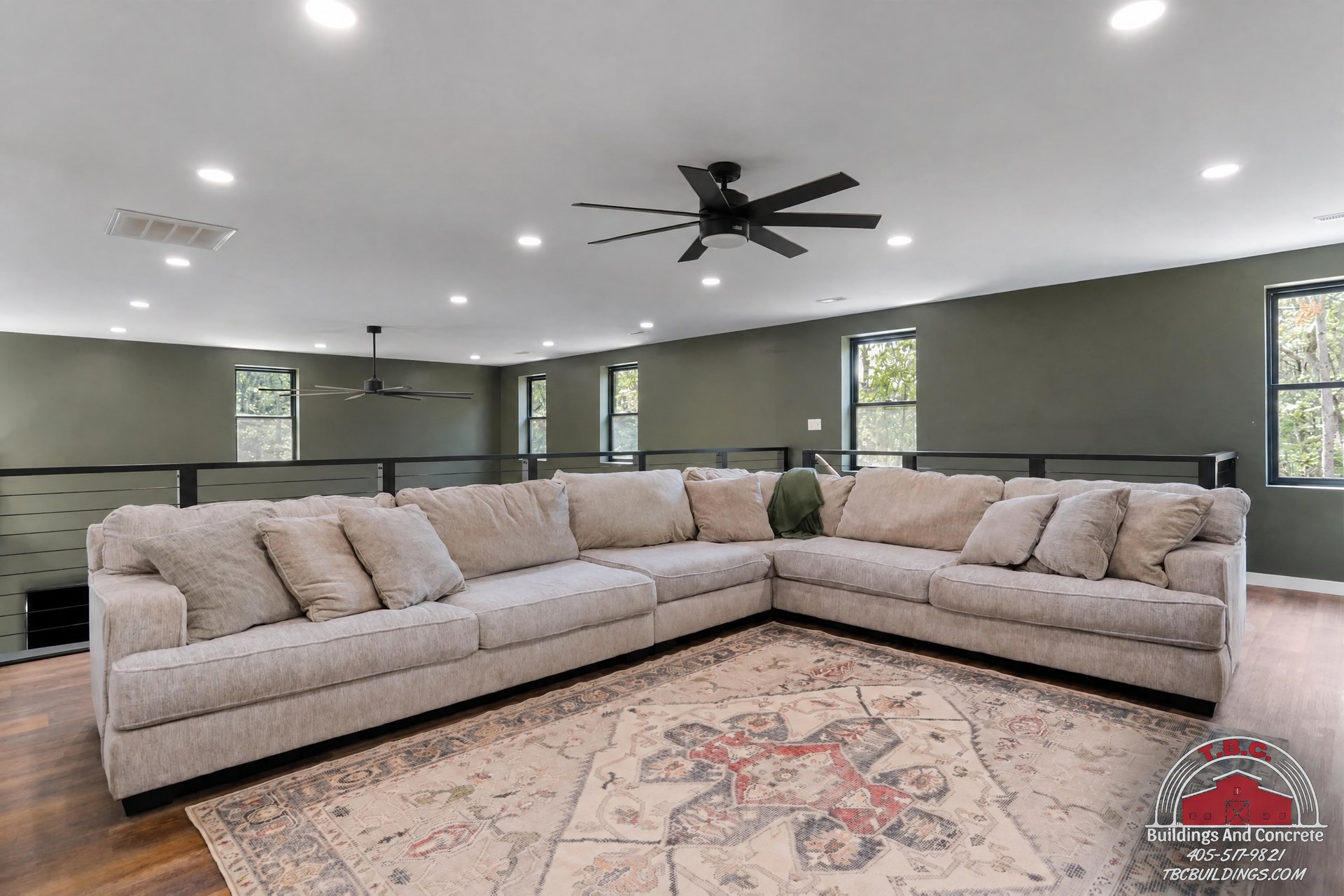 TBC project photo of a great room with a large sectional sofa, patterned area rug, ceiling fan, and black railing.