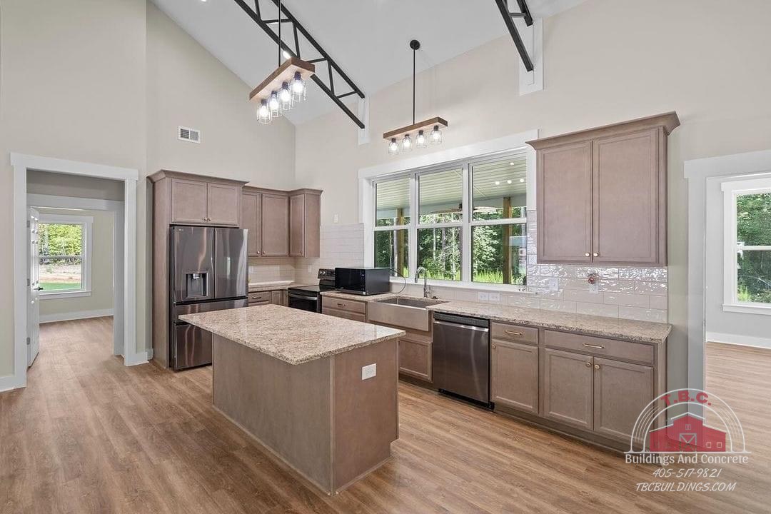 TBC project photo of a kitchen with wood flooring, an island, pale cabinets, stainless steel appliances, and large windows.