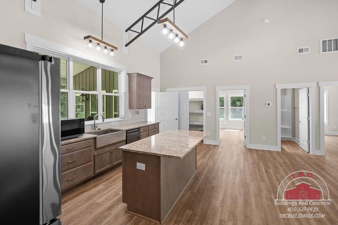 TBC project photo of a kitchen with an island, pale cabinets, wood flooring, and exposed ceiling trusses.
