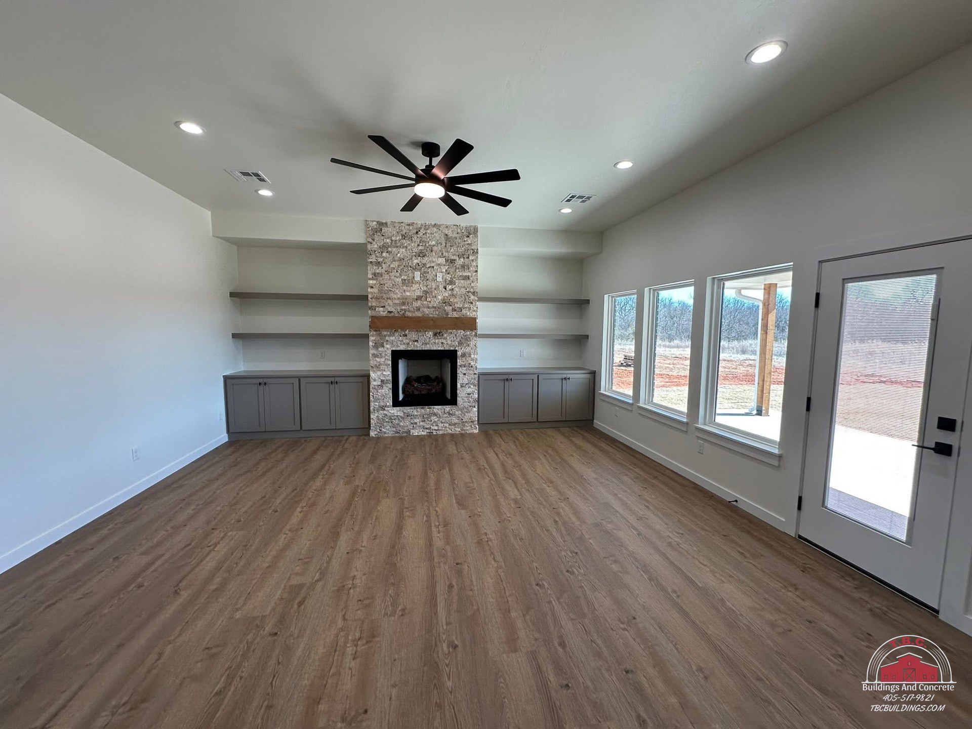 TBC project photo of a great room with wood flooring, a white shiplap fireplace wall, a ceiling fan, and large windows.