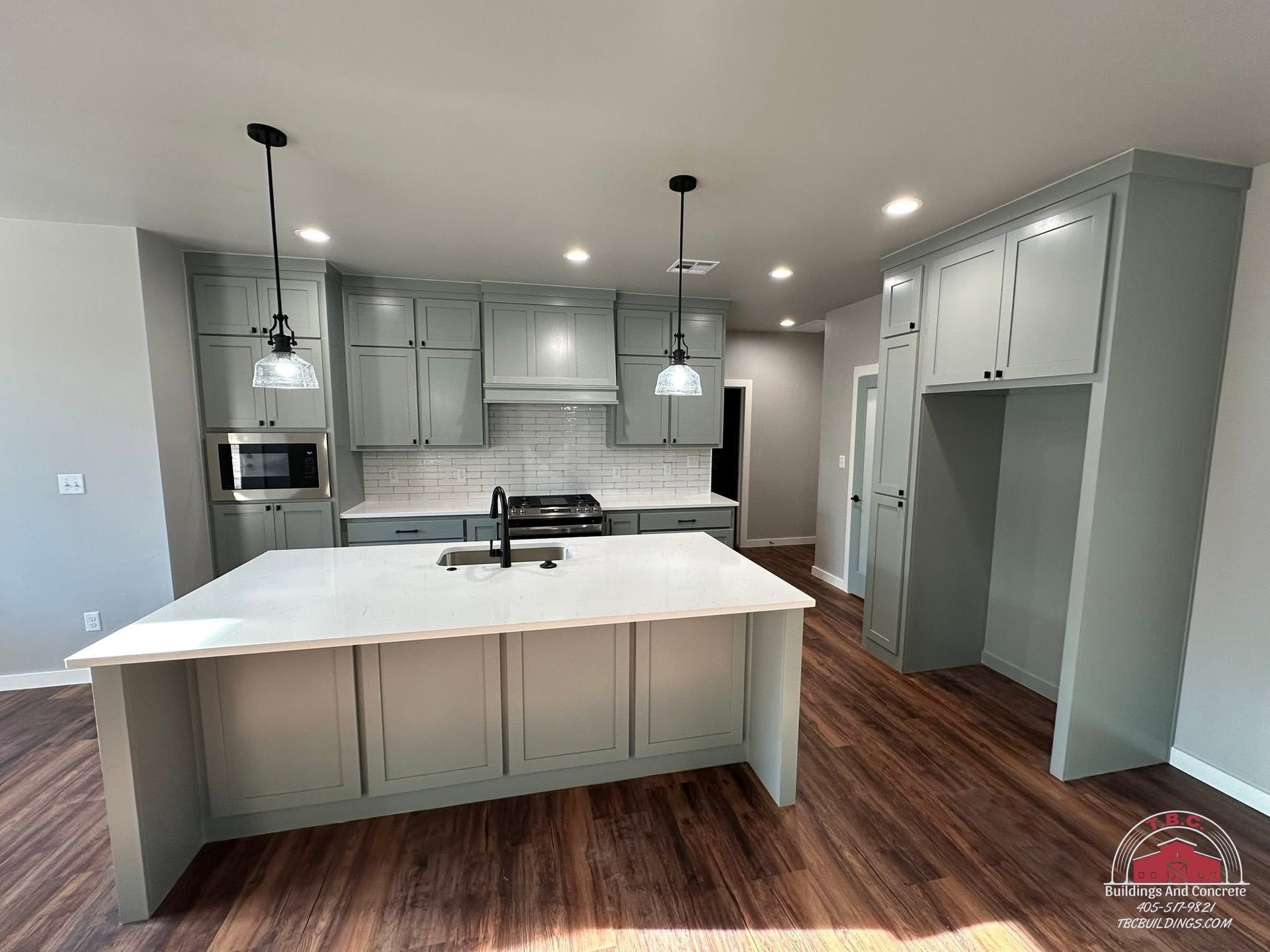 TBC project photo of a kitchen with a white island, gray cabinets, pendant lights, and wood flooring.