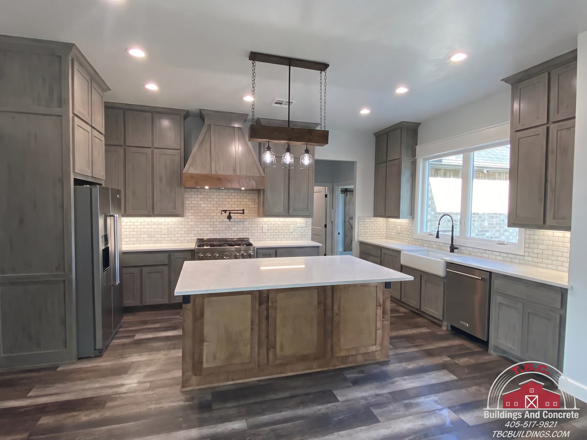 TBC project photo of a kitchen with gray cabinets, a central island, pendant lights, and wood flooring.