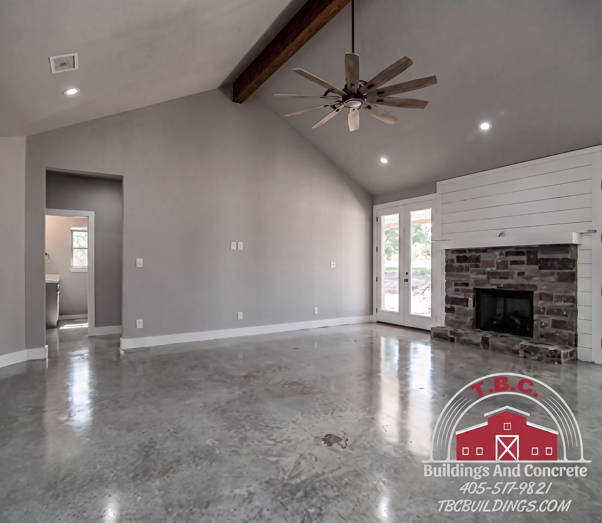 TBC project photo of an open great room with a white brick fireplace, ceiling fan, and large windows.