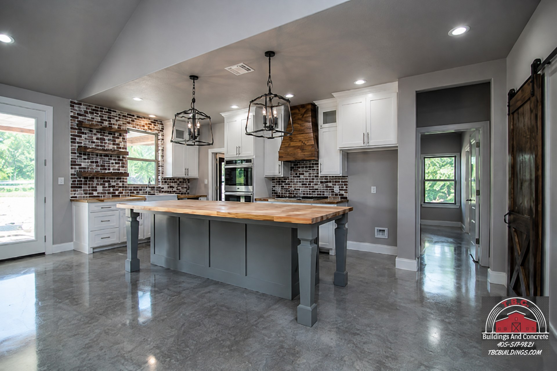 TBC project photo of an open kitchen with a wood-topped island, pendant lights, brick accents, and polished concrete flooring.