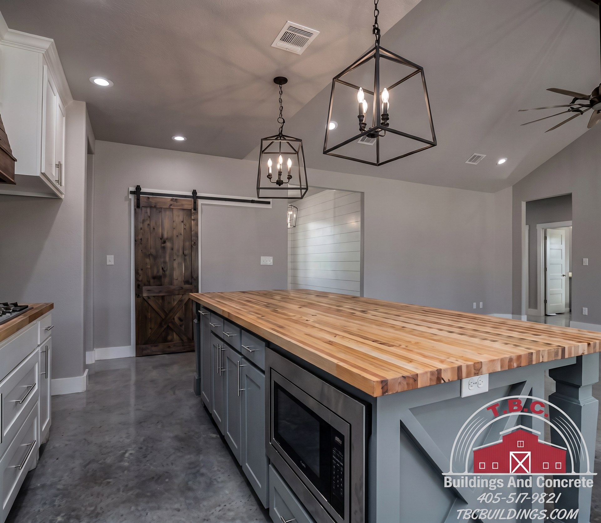 TBC project photo of a kitchen with a long wood-topped island, pendant lights, white cabinets, and polished concrete flooring.
