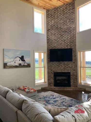 TBC project photo of a living room with a gray sectional, stone fireplace wall, and wood-plank ceiling.