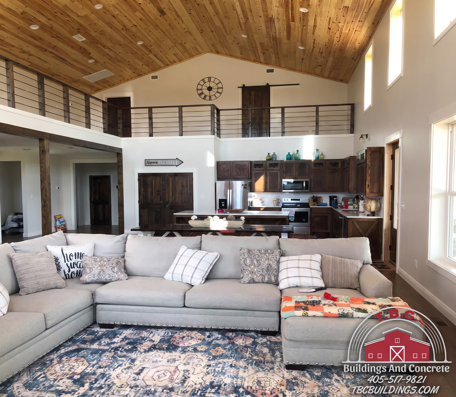 TBC project photo of an open great room with sectional seating, a kitchen island, upper loft railing, and wood ceiling.