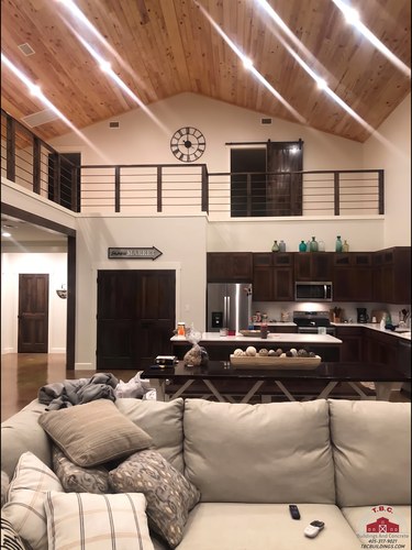 TBC project photo of a living room with sectional seating, a fireplace, loft railing, and wood ceiling.