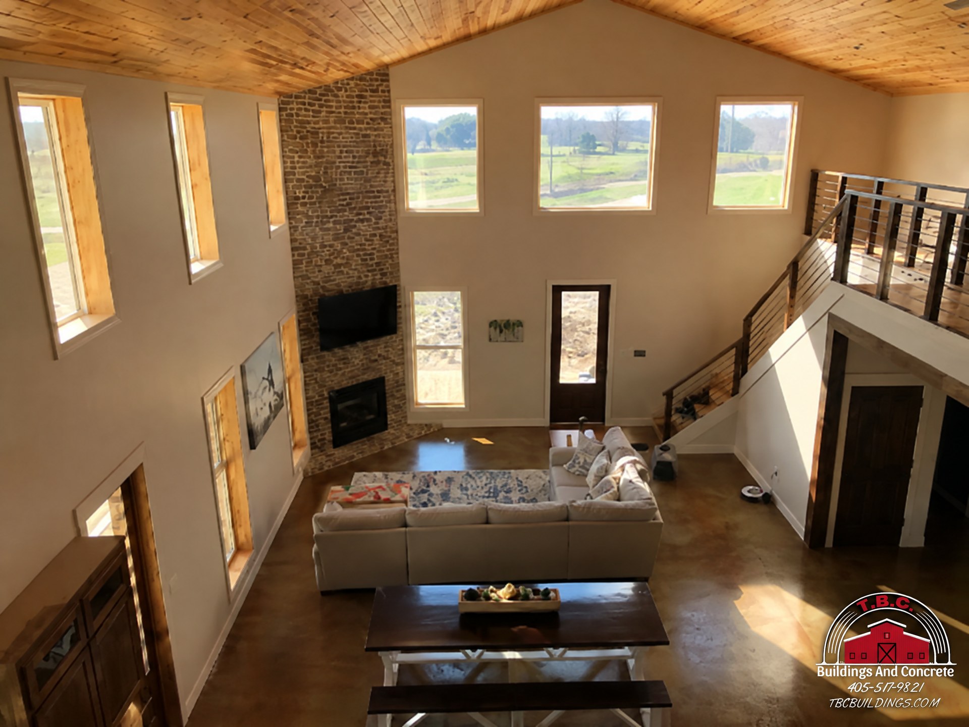 TBC project photo of a double-height great room with a fireplace, sofa seating, staircase, and upper railing.
