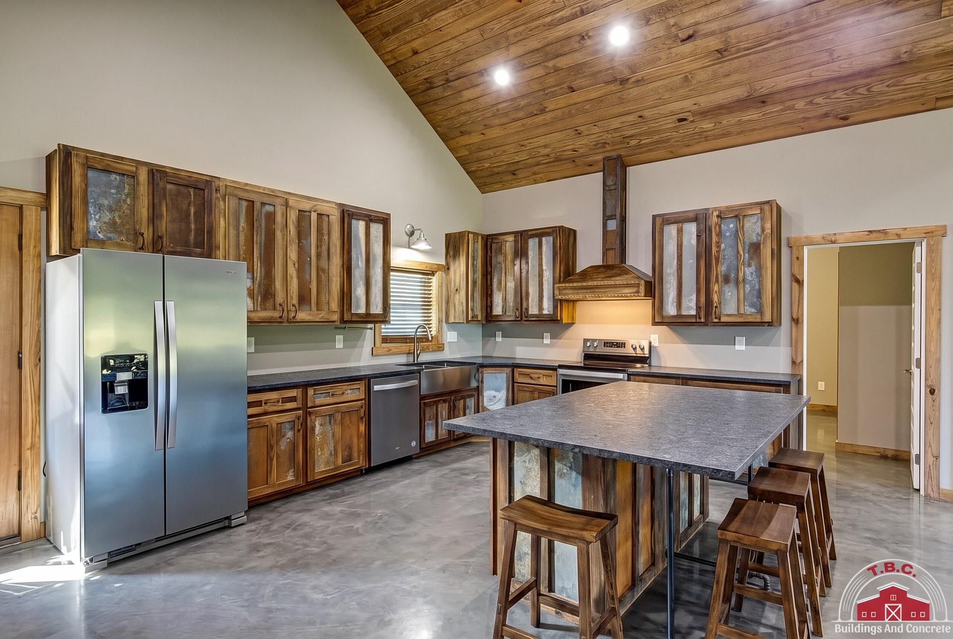 TBC project photo of a kitchen with wood cabinetry, stainless refrigerator, center island, and vaulted wood ceiling.